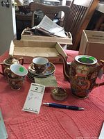Tea set displayed on a table including teapot, sugar bowl, creamer, cups, saucers, and small dishes with a handwritten note and a pen for scale.