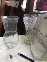 Photo showing three glass vases on a white surface. One is a clear, geometric vase, another is a cut glass vase with decorative patterns, and the third is a simple tall clear glass vase.