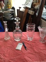 Four pressed glass items displayed on a pink cloth showing varying sizes and cut patterns, including decanter with stopper and three vases.
