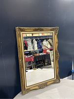 Full front angled view of large wall mirror showing gold tone ornate frame and reflective glass surface with items reflected in background.