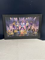 Frontal image of 'Legends Theatre' art print on board with black metal frame leaning against a dark blue wall, showing full artwork and frame.