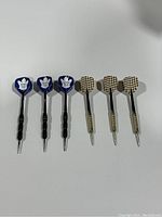 Six vintage darts arranged side by side: 3 darts with blue flights featuring white maple leaf emblem and black metal shafts; 3 darts with black and white checkered flights and brass-colored shafts.