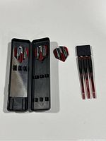 Three darts with flights next to an open black plastic case with metal panel inside, showing slots for parts