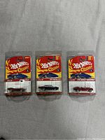 Three Hot Wheels Classics diecast cars in original blister packaging laid on grey fabric background. Includes silver 1967 Pontiac GTO, black 69 Camaro, and maroon 1970 Chevelle Convertible.
