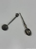 Two detailed vintage silver teaspoons with distinct decorative engravings and figures, showing front and side views.