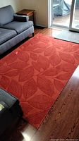 Full view of the orange-red area rug with leaf pattern laid on wood floor in living area next to grey couch and sliding glass door