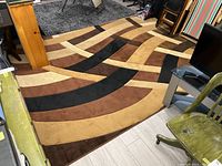 Area rug laid out on floor, showing abstract multi-colored pattern with beige, brown, black, and tan bands creating a geometric design.