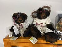 Two Danychuk Collection bears seated on a wooden surface, both in white dresses with burgundy ribbons.