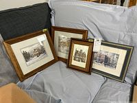 Four framed prints resting on gray and dark cloth surfaces. Wood frames with glass. One has a dark stain and the others are medium tone woods.