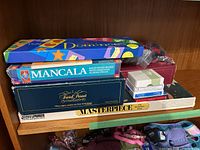 Photo showing stacked board games including Dominoes, Mancala, Trivial Pursuit, Masterpiece, some playing card decks, and game components in plastic bag
