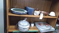 Photo of wooden shelf holding two Sango plates, three ceramic mugs, white egg coddler, glass deep dish round plate with lid and handle, Corning Ware style pots, and glass tray in original box.