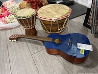 Blue Harmony youth guitar with missing strings and wear, laying flat with two wooden drums behind it.