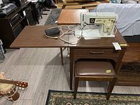 Wide photo of Singer Stylist sewing machine installed in laminate wood table with pull-out leaf extension and matching upholstered wooden stool beneath.