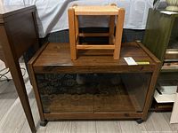 Wooden stool with woven twine seat placed on top of dark wood laminate media stand with glass doors and casters.