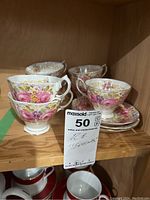 Eight teacups and three saucers from the Royal Albert Serena pattern displayed on a wooden shelf showing the floral design and some wear on gold edging.