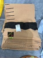 Folded tan wool blanket with black stripe detail, displayed to show full blanket size label and stitching.