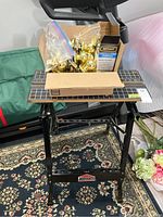 Jobmate folding work station with black metal base and composite top with measurement grids and angles. On top sits a cardboard box and plastic bags full of brass door hardware including knob sets and hinges.