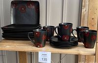 Set of black square plates and matching mugs with red floral designs displayed on wood shelf.