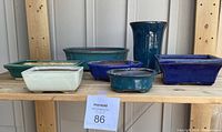 Full view of seven bonsai pots on wooden shelf, various colors and shapes, with auction lot tag visible.