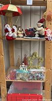 Photo showing various Christmas plush figurines including Santas with parachute umbrellas and snowmen on shelves above a red lidded tote and assorted small decorative trees and garlands below.