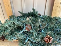 Close-up showing green artificial pine needles, embedded string lights with green wiring, and brown pinecones attached.