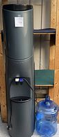 Full view of tall cylindrical gray water cooler with a large blue water bottle next to it, showing size and setup.