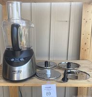 Full view of Black & Decker PowerPro food processor base, food bowl, and three cutting blade discs with one blade adapter piece on wooden shelf.