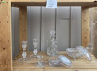 Full frontal view of the glassware items arranged on a wooden shelf showing one decanter, three assorted sized candle holders, a covered dish, and two serving dishes or bowls.
