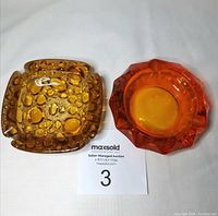 Top and angled top view of amber pebbled glass ashtray and bright orange diamond-patterned edge glass ashtray side by side on white background with auction card.