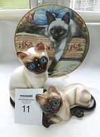 Photo showing the two Siamese cat figurines with blue eyes positioned in front of the Franklin Mint collector plate depicting a Siamese cat on a patterned cushion.