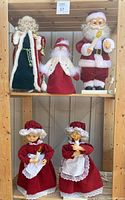 Five vintage animated Christmas figures arranged on wooden shelves, two Santa Clauses on top shelf with a smaller Santa in the middle, two Mrs. Claus figures on bottom shelf dressed in matching red dresses holding candles.
