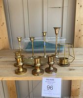 Overview of 8 brass candle holders of various styles and sizes placed on wooden surface with plastic container in background.