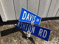Two blue rectangular metal street signs, one reading 'DAVIS DR' and the other 'ASHTON RD,' connected by a metal bracket, laying on ground near a building.