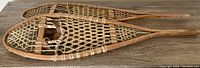 Pair of large antique snowshoes laid flat on wooden surface showing full length, wood frame, rawhide webbing and leather toe straps.