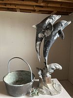 Photo showing tall cast metal dolphins sculpture with two dolphins jumping over stylized waves, antique witches cauldron to side, and small frog spigot at base.