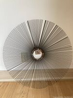 Front view of large metal starburst wall sculpture showing thin metal rods radiating symmetrically from a central circular ring, placed on wood floor against white wall.