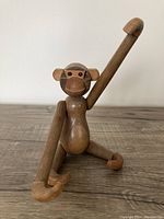 Frontal view of the wooden jointed monkey toy showing raised arm and seated pose on wooden surface, highlighting wood grain and articulated joints.