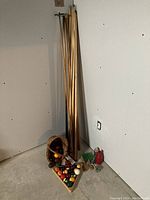 Photo showing the 12 pool cues leaning against the wall with the basket and triangle rack containing billiard balls on the floor, accompanied by small accessories.