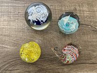 Overview photo showing all four Murano glass paperweights on wooden surface: white feather motif sphere, seagull motif sphere, yellow speckled dome, and snail-shaped paperweight.