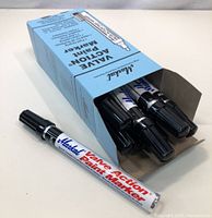 Box of 12 Markal Valve Action Paint Markers, black ink, with markers partially visible inside box and one marker out on table.