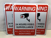 Three metal surveillance signs stacked leaning against a wall, showing front.