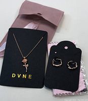 Necklace and earrings displayed on black cards with brand name DVNE, accompanied by a pink fabric pouch.
