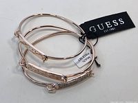 Three new Guess bracelets with tags, shown in stacked arrangement, featuring rose gold-tone metal and different decorative plates.