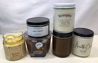 Six assorted new candles in glass jars of varying sizes and colors. One candle is yellow with lavender buds and missing lid and stone.