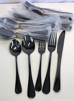 Photo showing 5 individual pieces of black cutlery (tablespoon, teaspoon, fork, dessert fork, knife) laid flat with bundles of identical black cutlery wrapped in plastic in the background.