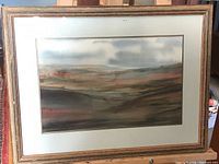 Framed watercolor landscape painting with a wooden frame and glass covering showing a muted color palette of a scenic landscape.