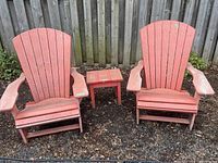 Pair of red resin Muskoka chairs and matching red table outside on ground with fence background. Showing weathered surface on all items.