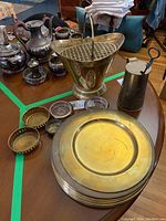 Photo showing multiple metal charger plates stacked, silver plate coasters, vintage brass smudge pot fire starter, and other metal items on a wooden table.