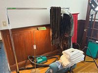 Metal clothing rack with horizontal bar, set against wooden wall paneling. Some clothing items hanging and other unrelated household items visible around but not included.