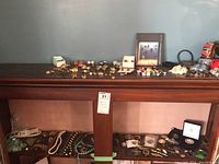 Two dark wood shelves and tabletop with assorted costume jewelry pieces spread out and partially boxed, including necklaces, bracelets, earrings, and brooches.